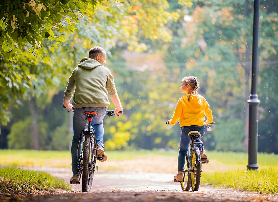 About Our Agency - Rear View of a Father and Daughter Riding Bikes in the Park on a Sunny Day